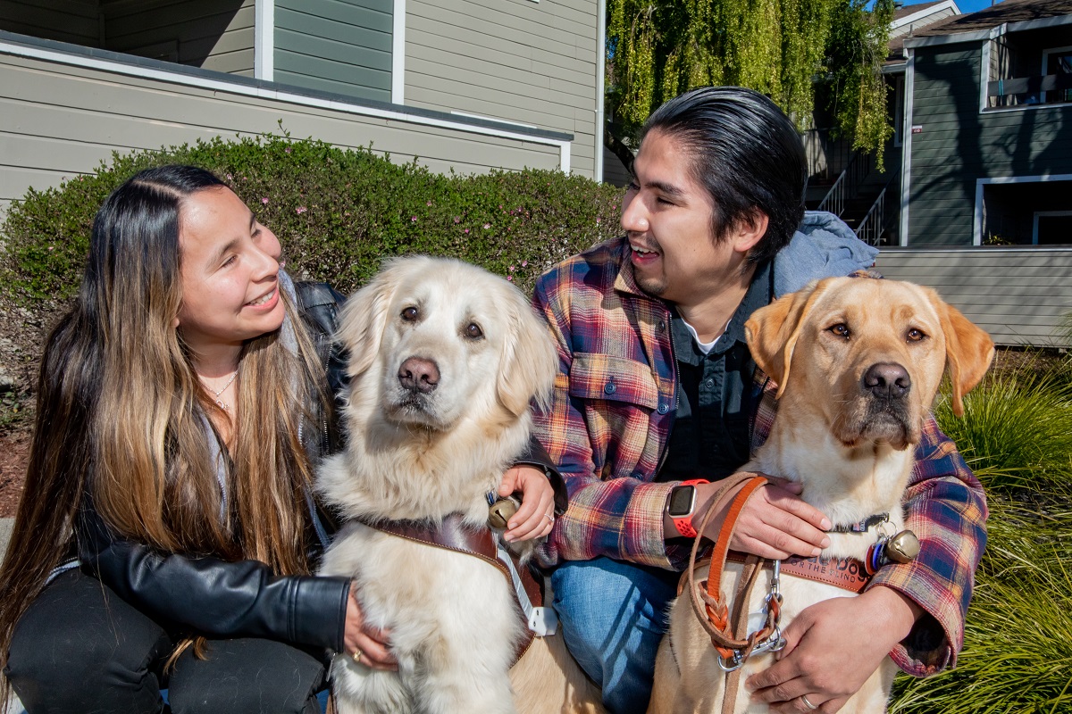 Marissa and Alex smile at one another while sitting with their guide dogs, a Golden Retriever and a golden Lab.